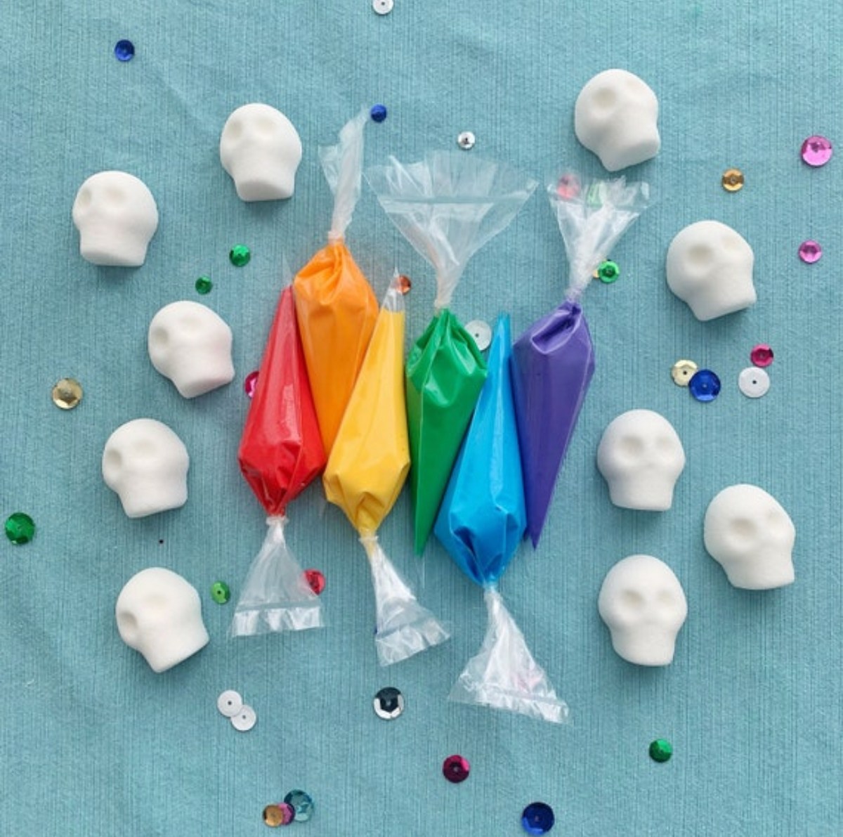 Royal icing bags in colors red, orange, yellow, green, blue and purple lined up in a row, surrounded by 10 small sugar skull cubes and sequins scattered lightly throughout the photo frame.