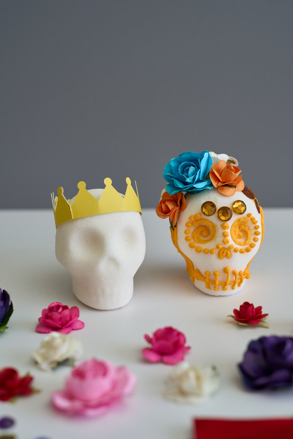 A white decorative skull centerpiece on a white surface, decorated with a yellow paper crown on the head and blue and orange paper flowers on top. A blank white sugar skull head wearing only the yellow crown stands beside it.