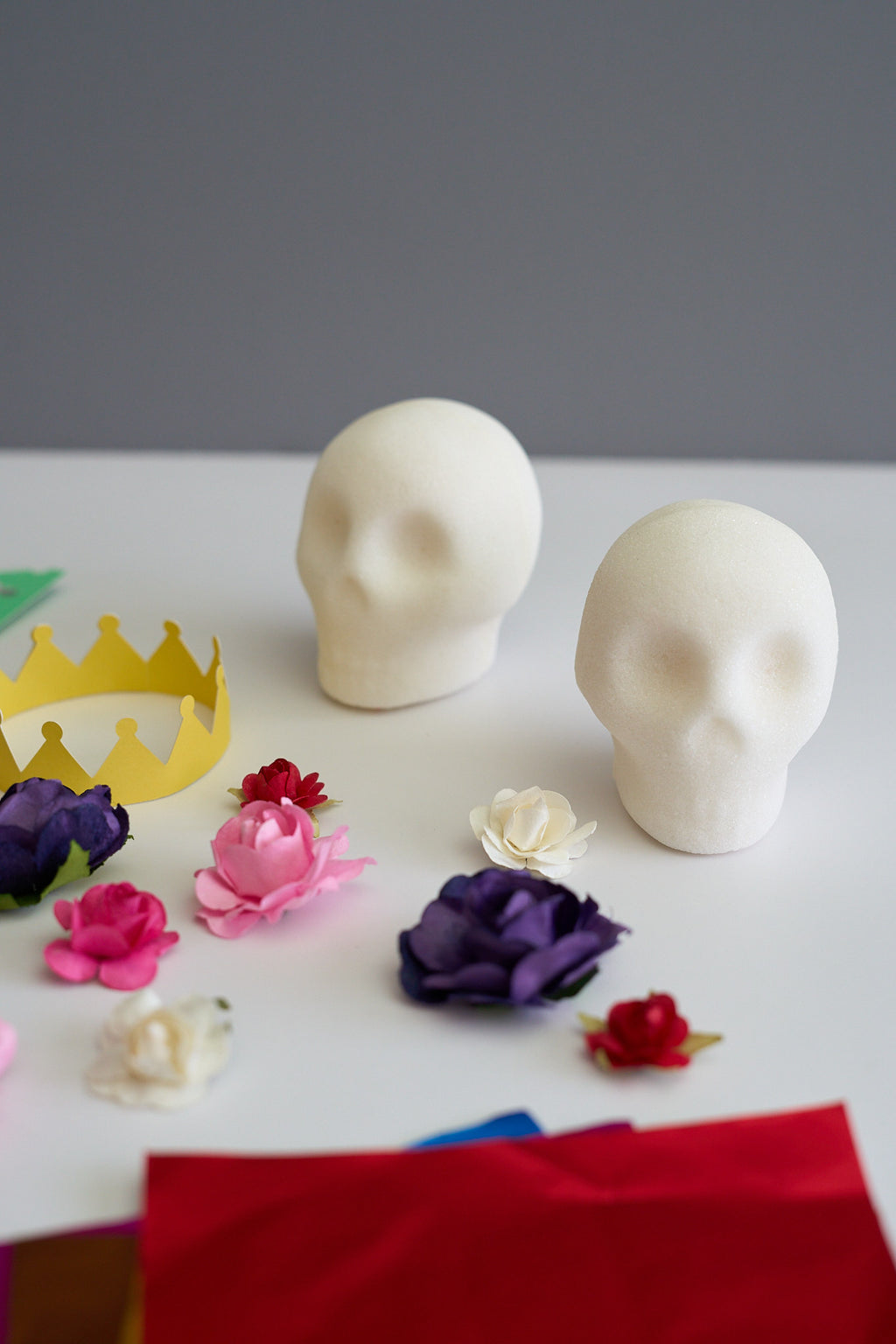 Two blank white skulls made of sugar on a white table with colorful paper flowers, a yellow crown, and red paper sheets against a gray background.