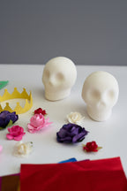Two blank white skulls made of sugar on a white table with colorful paper flowers, a yellow crown, and red paper sheets against a gray background.