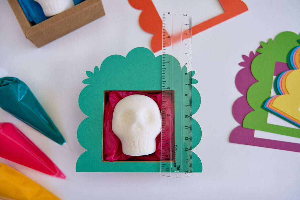 A white skull-shaped item displayed inside a green scalloped box with pink tissue paper, alongside a clear ruler for scale, on a white surface with colorful craft supplies around it.