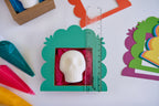 A white skull-shaped item displayed inside a green scalloped box with pink tissue paper, alongside a clear ruler for scale, on a white surface with colorful craft supplies around it.