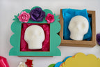 Two white skull blanks displayed in decorative gift boxes on a white surface. One is in a green scalloped frame with pink tissue paper and paper flowers, and the other is in a brown kraft box with blue tissue paper.
