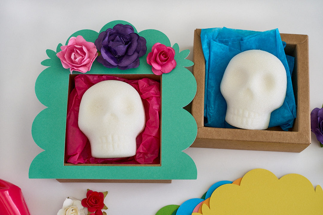 Two white skull blanks displayed in decorative gift boxes on a white surface. One is in a green scalloped frame with pink tissue paper and paper flowers, and the other is in a brown kraft box with blue tissue paper.