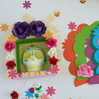 A colorful handmade Day of the Dead (Dia de los Muertos) decorative frame on a white surface, shown alongside a matching multi-layered paper cutout frame. The frame features purple paper roses, pink and red fabric flowers, a white sugar skull decorated with blue and yellow details, and gold sequins around the border.