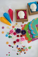 A colorful craft or gift set arranged on a white surface, including two white skull-shaped items in brown boxes with pink tissue paper, three piping bags in yellow, blue, and pink, scattered flower and star embellishments, sequins, and a stack of bright paper cutouts.