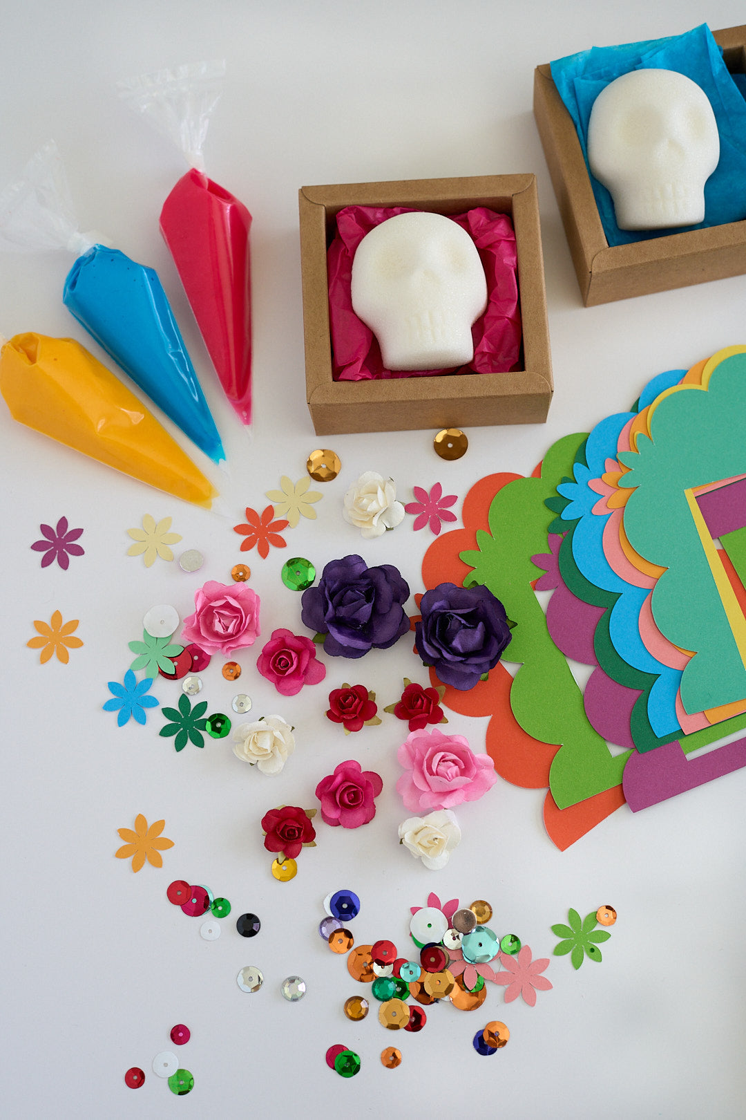 A colorful craft or gift set arranged on a white surface, including two white skull-shaped items in brown boxes with pink tissue paper, three piping bags in yellow, blue, and pink, scattered flower and star embellishments, sequins, and a stack of bright paper cutouts.