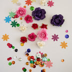 A flat-lay arrangement of craft supplies on a white background, including artificial rose-shaped embellishments in pink, purple, red, and white, paper flower cutouts in multiple colors, and scattered round sequins.