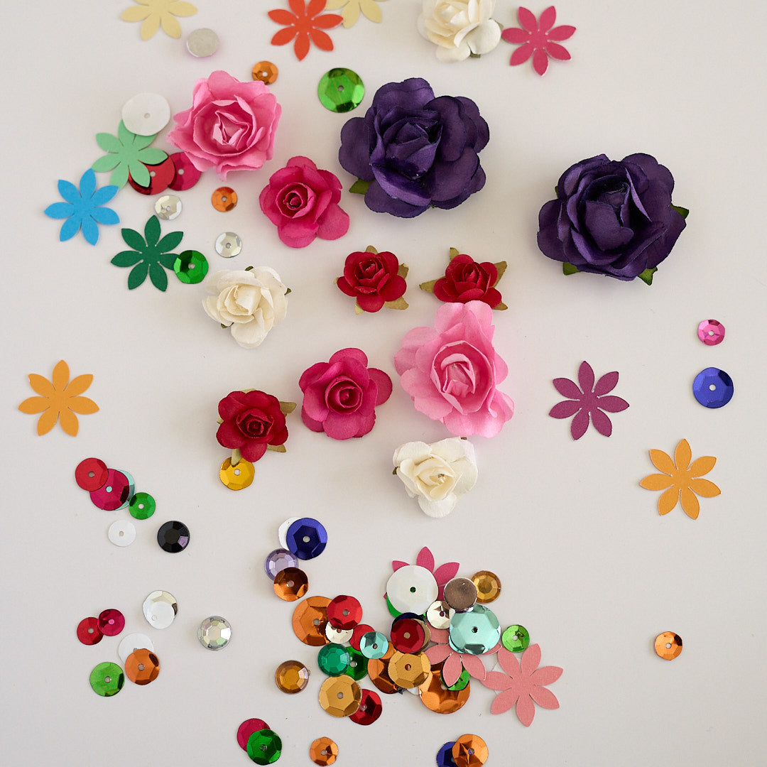 A flat-lay arrangement of craft supplies on a white background, including artificial rose-shaped embellishments in pink, purple, red, and white, paper flower cutouts in multiple colors, and scattered round sequins.