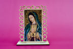 A small laser cut wooden devotional plaque featuring a painted image of Our Lady of Guadalupe, set in an ornate openwork wooden frame and displayed on a laser cut base against a solid pink background.