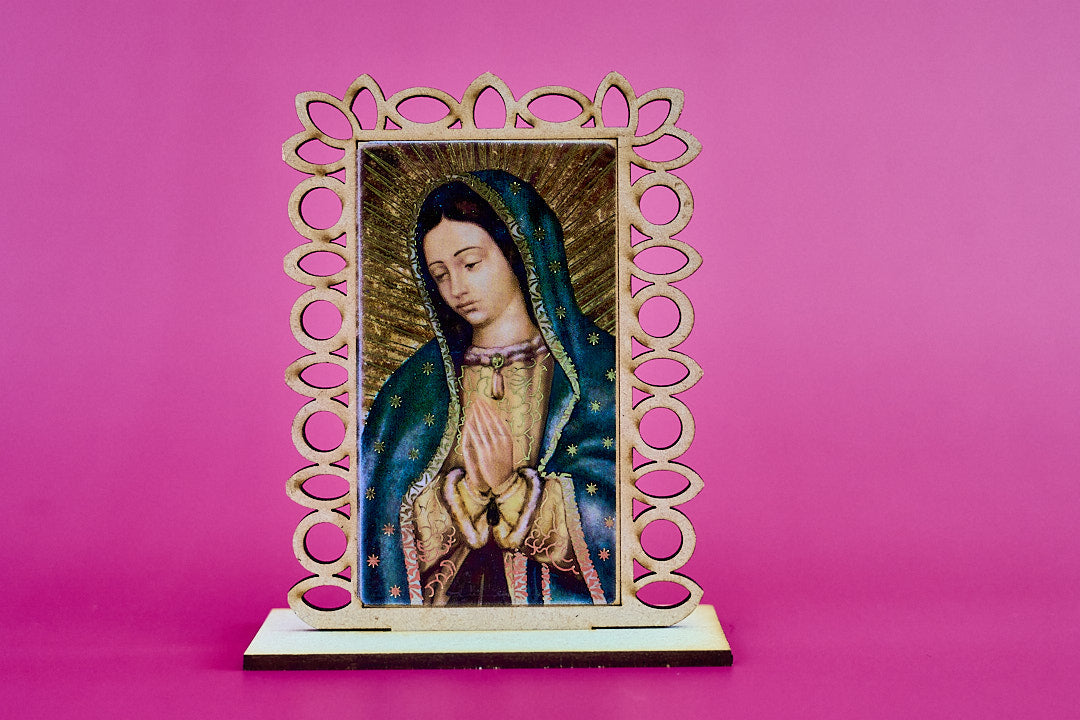 A small laser cut wooden devotional plaque featuring a painted image of Our Lady of Guadalupe, set in an ornate openwork wooden frame and displayed on a laser cut base against a solid pink background.