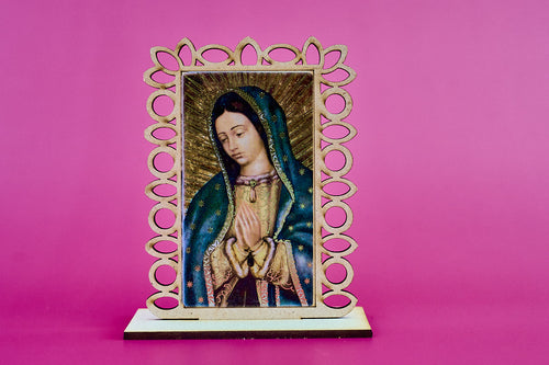 A small laser cut wooden devotional plaque featuring a painted image of Our Lady of Guadalupe, set in an ornate openwork wooden frame and displayed on a laser cut base against a solid pink background.