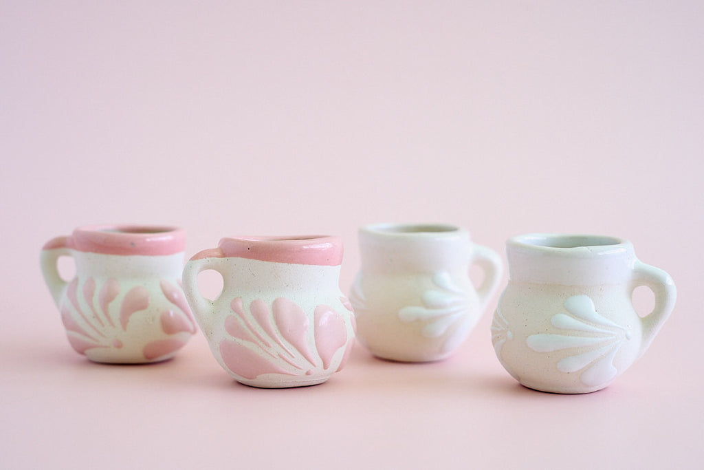Four small ceramic cups with side handles, decorated with embossed floral patterns in pale pink and white, arranged in a row against a soft pink background.