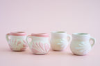Four small ceramic cups with side handles, decorated with embossed floral patterns in pale pink and white, arranged in a row against a soft pink background.