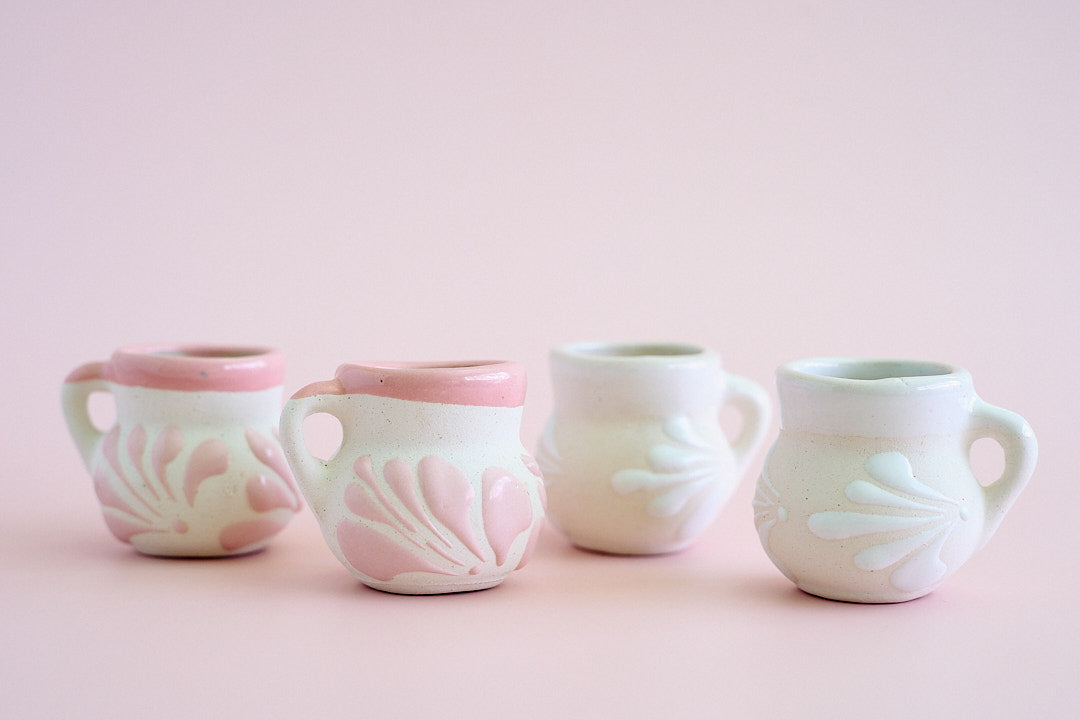 Four small ceramic cups with side handles, decorated with embossed floral patterns in pale pink and white, arranged in a row against a soft pink background.