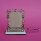 A laser-cut decorative frame with a scalloped, circular cutout border in a light wood tone, shown standing upright with a base and a clear plastic ruler scale against a solid pink background.