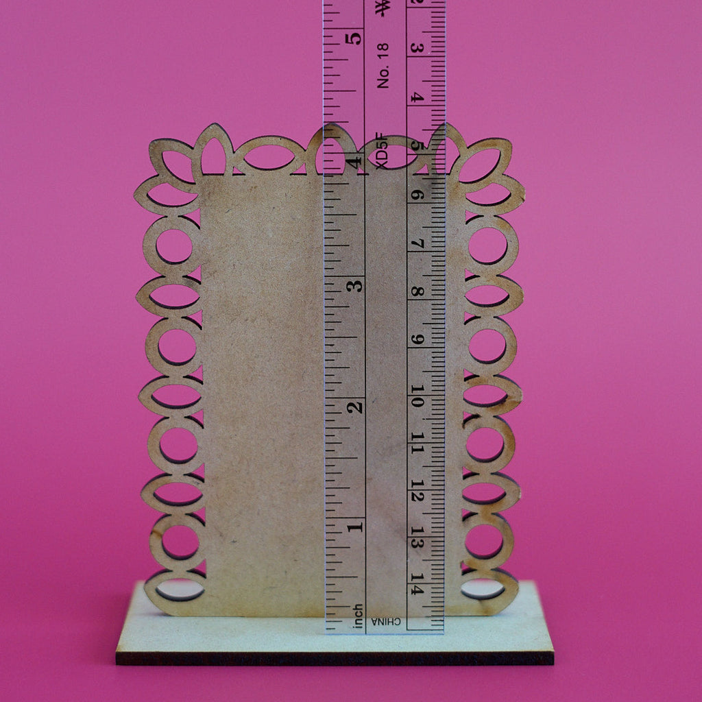 A decorative wooden frame or sign holder with an ornate scalloped edge and circular cutouts, shown on a white base with a ruler standing vertically to indicate dimensions, against a solid pink background.