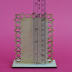 A decorative wooden frame or sign holder with an ornate scalloped edge and circular cutouts, shown on a white base with a ruler standing vertically to indicate dimensions, against a solid pink background.