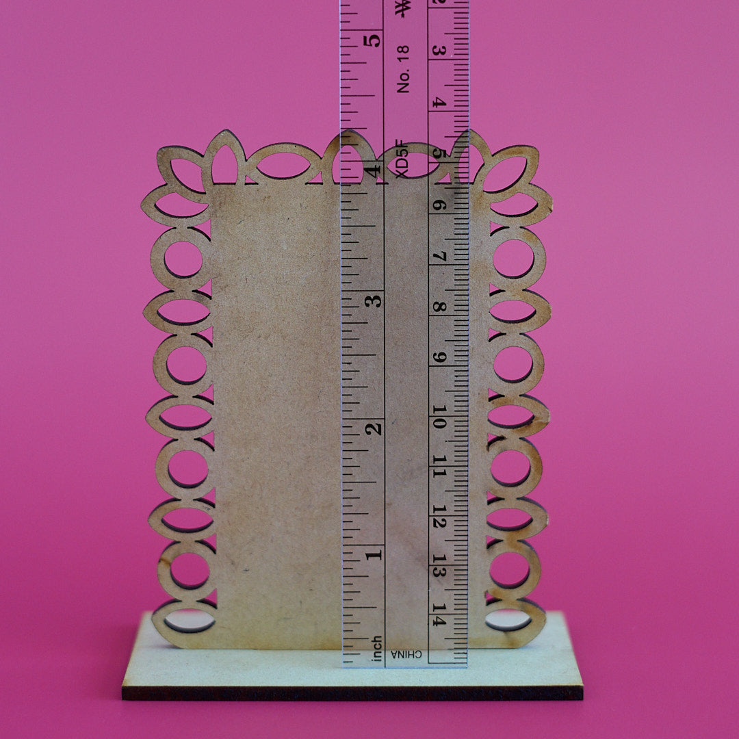 A decorative wooden frame or sign holder with an ornate scalloped edge and circular cutouts, shown on a white base with a ruler standing vertically to indicate dimensions, against a solid pink background.