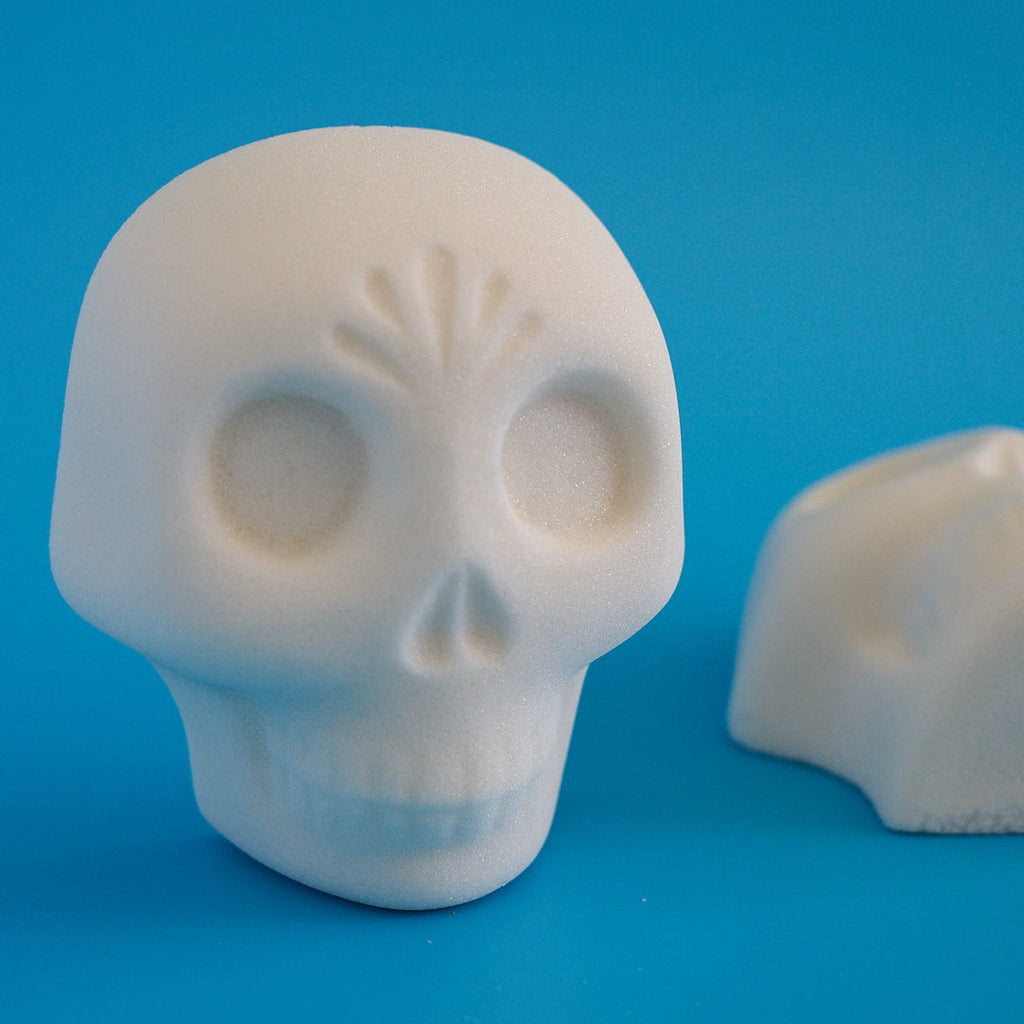 A white sugar skull figurine with Day of the Dead style engravings, shown on a vibrant blue background with a second skull partially visible in the background.