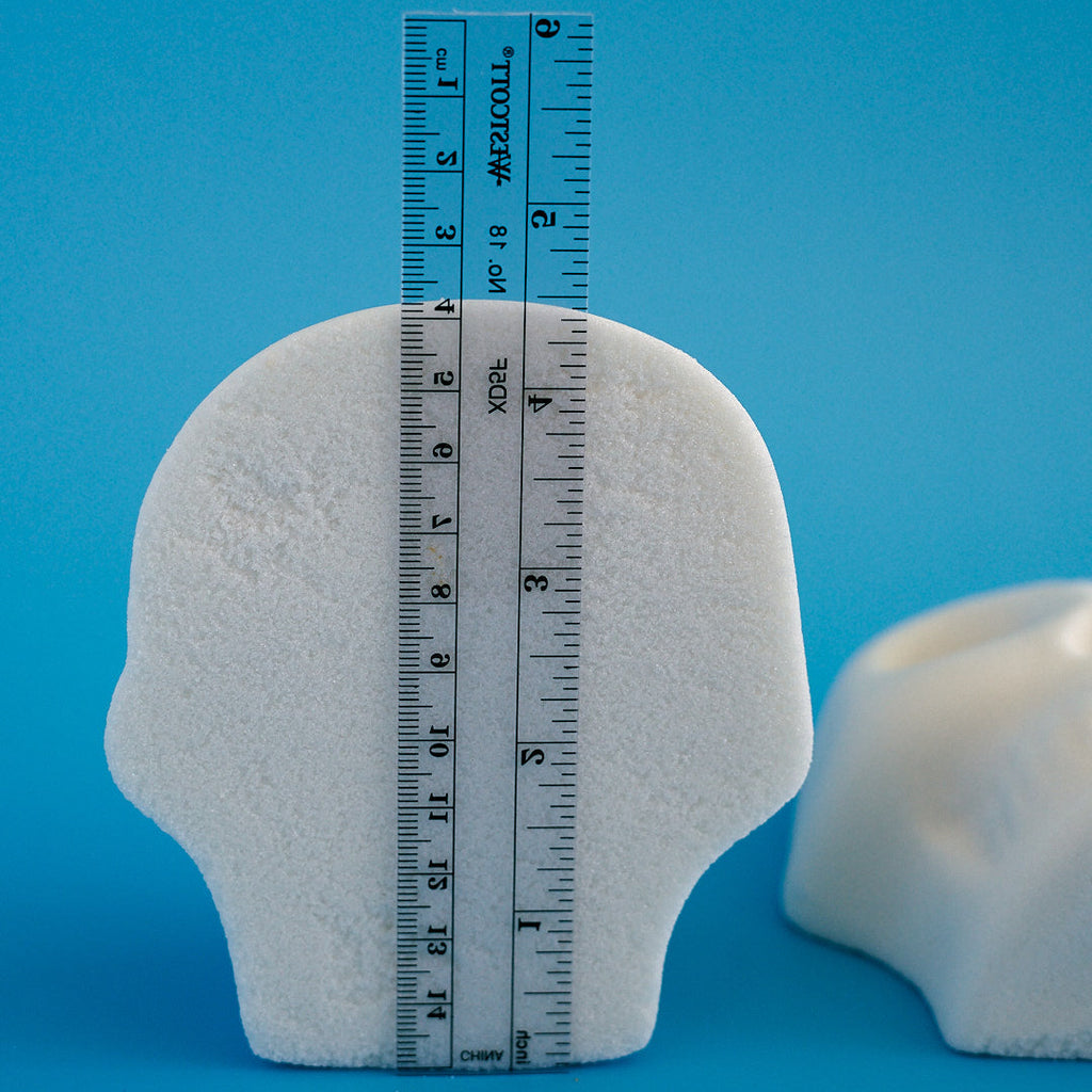 A white flat back head-shaped object stands upright against a bright blue background, with a black measuring ruler placed vertically in front of it. A second similar white object is partially visible on the right edge laying down.