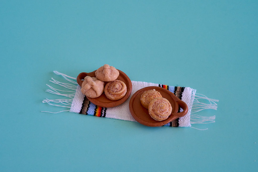 Two small brown ceramic plates holding miniature pastries, arranged on a white woven textile with colorful stripes and fringe, set against a solid turquoise background.