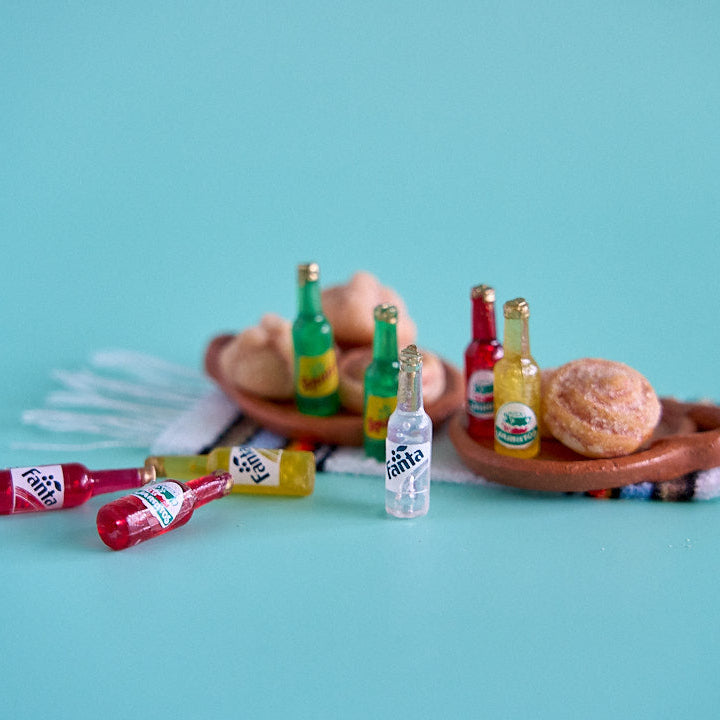 A miniature diorama setup featuring tiny toy beverage bottles and donuts on a terracotta tray, set against a solid turquoise background.