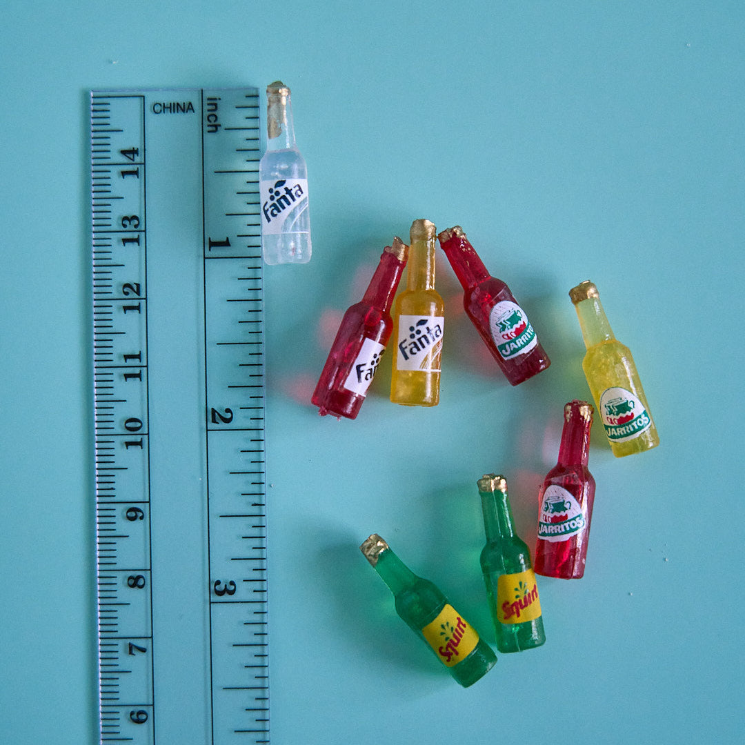 A set of miniature Fanta soda bottle charms arranged next to a clear plastic ruler on a solid light blue background.