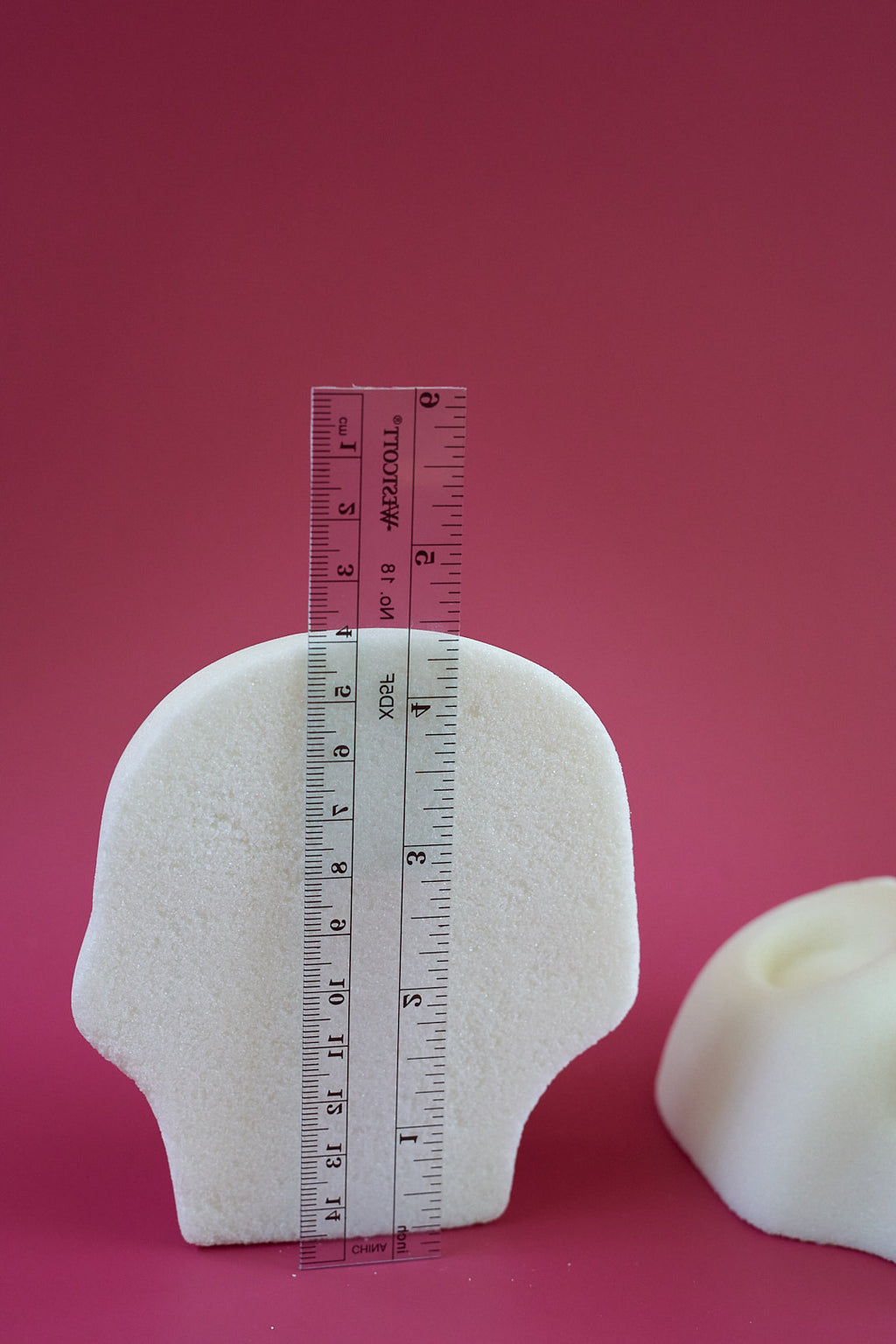 A white blank sugar skull standing upright on a pink background, with a transparent ruler placed vertically against it to show height. A second similar object is partially visible on the right.