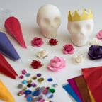 A flat-lay of craft or party decoration supplies on a white surface, including two white sugar skull blanks (one with a yellow paper crown), colorful piping bags, small paper flowers, and scattered rhinestones.
