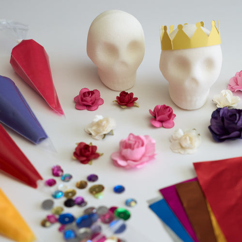 A flat-lay of craft or party decoration supplies on a white surface, including two white sugar skull blanks (one with a yellow paper crown), colorful piping bags, small paper flowers, and scattered rhinestones.