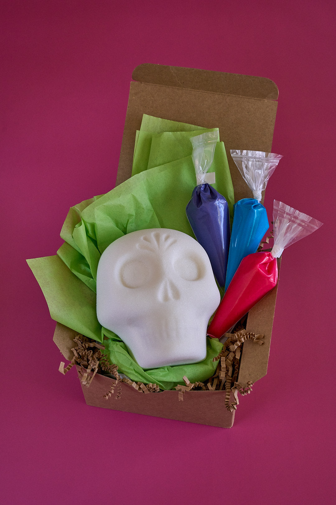 A white skull-shaped blank sugar skull made of sugar displayed in a brown gift box with green tissue paper and three colorful piping bags in purple, blue, and red, set against a solid magenta background.