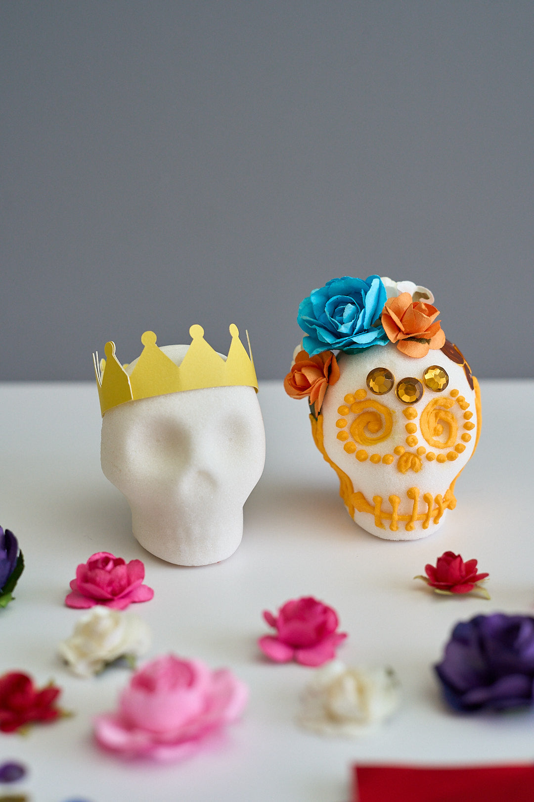 A decorative sugar skull centerpiece on a white surface against a gray background, featuring a white skull with orange and blue paper flowers on its head and a yellow crown on a second white blank sugar skull head. 