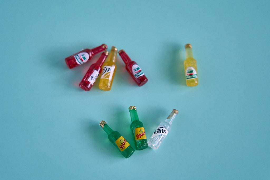 Six miniature Fanta soda bottles in red, yellow, green, and clear plastic, arranged on a solid light blue background.
