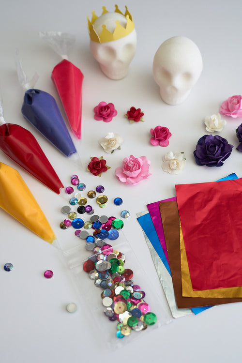 A flat-lay of craft supplies on a white background, including piping bags filled with red, purple, yellow, and pink icing, small paper roses in multiple colors, a clear bag of assorted rhinestones, sheets of red, gold, and metallic pink paper, and two white sugar skull blank heads wearing a yellow crown.